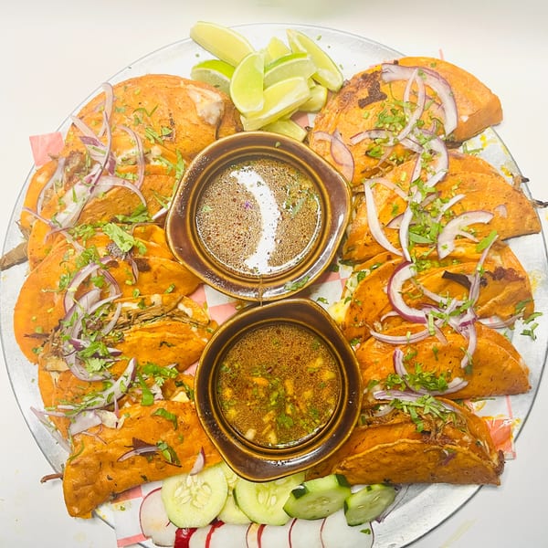 A platter of Mister Taco birria tacos with red onions, cilantro, lime, and two bowls of consommé.